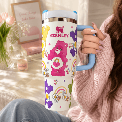 A person in a pink knit sweater holds a Stanley insulated tumbler with a white body, pastel multicolor bear and flower graphics, and a light blue handle. The background is a softly lit indoor setting with flowers and framed decor.