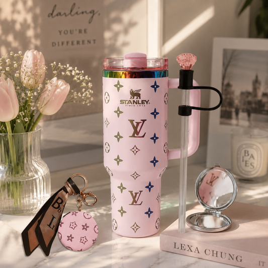 A pink Stanley tumbler with a Louis Vuitton-style monogram pattern is styled on a marble surface with a matching pink compact mirror, a pink keychain, and a clear straw accessory with a pink gem handle. The background features soft pink tulips in a glass vase and a framed print.
