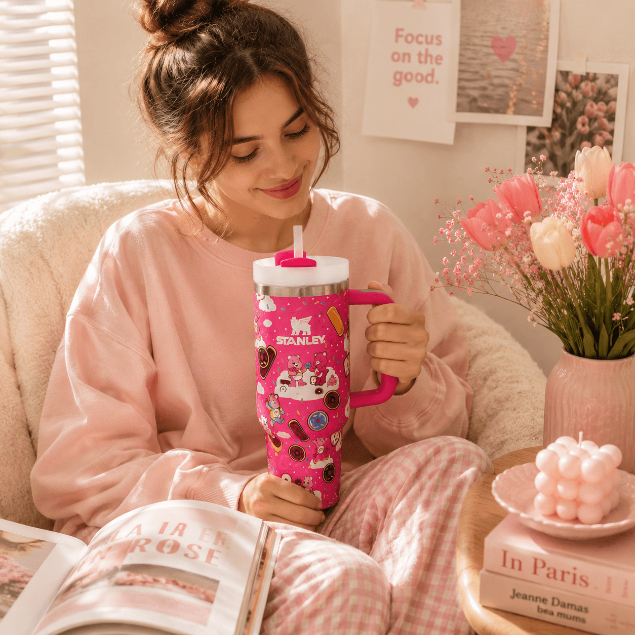 A woman in a pink lounge set holds a pink handled tumbler with a white lid and a straw. The tumbler is decorated with illustrations of a cat, donuts, and the text 'ST.