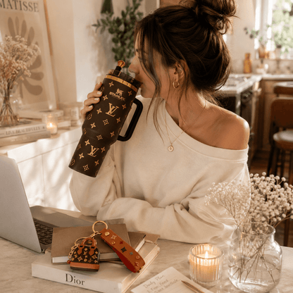 A woman in a cozy home setting drinking from a brown Louis Vuitton monogram water bottle, with a laptop, books, and candles on the table.