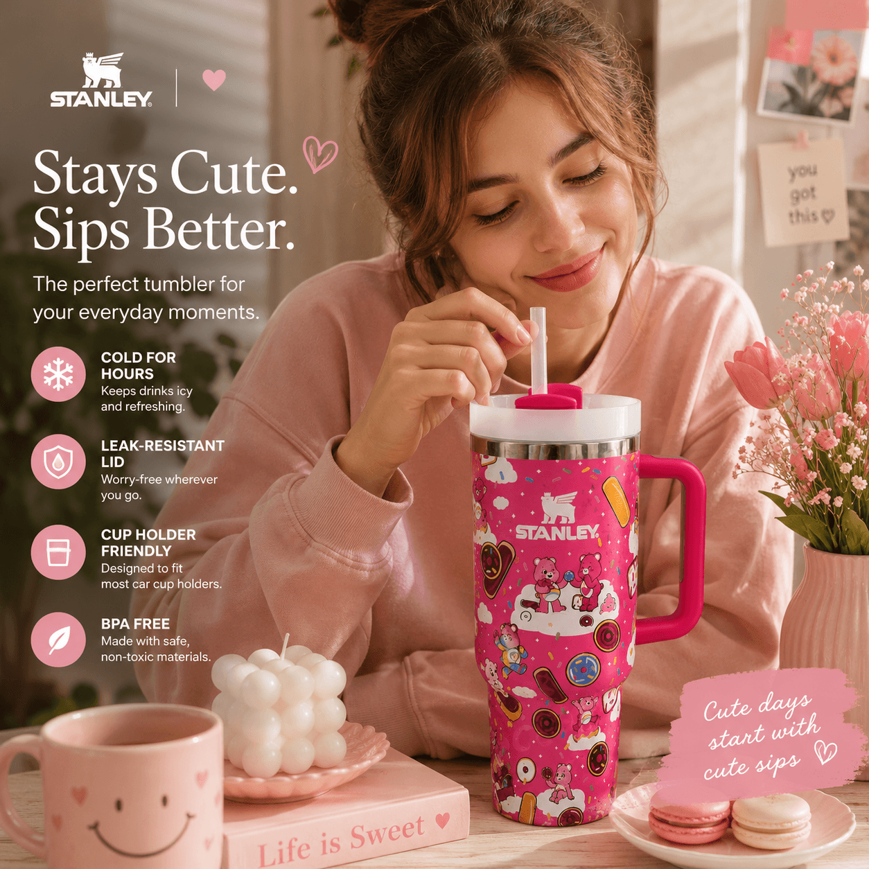 A woman in a pink sweatshirt holds a pink Stanley tumbler with a handle and straw, decorated with cartoon illustrations. The scene is a cozy indoor setting with flowers, macarons, and promotional text highlighting the tumbler's features.