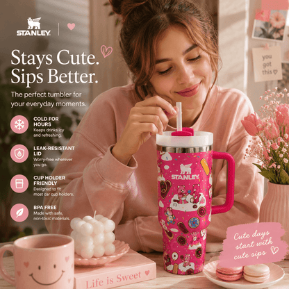A woman in a pink sweatshirt holds a pink Stanley tumbler with a handle and straw, decorated with cartoon illustrations. The scene is a cozy indoor setting with flowers, macarons, and promotional text highlighting the tumbler's features.