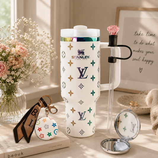 A white Stanley travel mug with a Louis Vuitton-inspired monogram pattern, styled on a tabletop with a clear glass tumbler, compact mirror, and a small round pouch in a bright, sunlit room.
